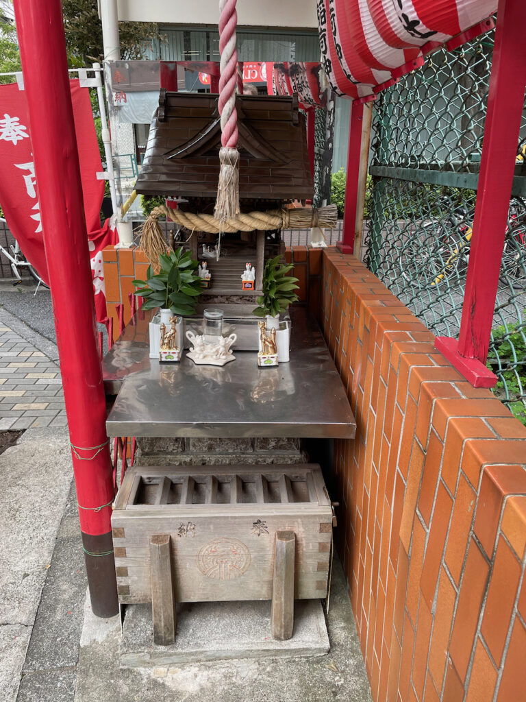 The sanctuary of the jinja, with an offering of water, sakaki, and ceramic foxes sitting on a metal table in front of it. There is even a bell rope, but it hangs from the roof over the sanctuary, not the sanctuary itself.