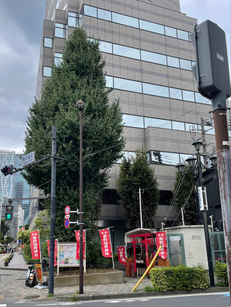 A small Inari jinja with a red torii behind a large single tree. Red banners around the tree make it clear that this tree is the sacred forest for the jinja.