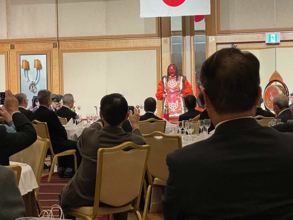 A masked dancer, in red, stands below a Japanese flag. People are seated at tables in the foreground, and some are taking photographs.